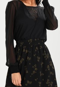Donna che indossa un abito nero con maniche lunghe trasparenti, dettagli in pizzo sul collo e una gonna a fiori con vita elasticizzata.