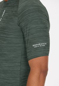 Dunkelgrünes Sportshirt mit einem strukturierten, gestreiften Muster. Beinhaltet weiße Markenkennzeichnung am Ärmel mit der Aufschrift "ENDURANCE HERO OF THE ROAD."