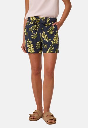 Donna che indossa pantaloncini blu navy con stampa floreale gialla, top bianco, sandali beige, in piedi con una mano in tasca.