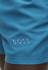 Μπλε υφασμάτινες βερμούδες με τη λέξη «BOSS» κεντημένη με αντίστοιχο μπλε νήμα κοντά στο τελείωμα, φορεμένες από άτομο με σκουρόχρωμο δέρμα.