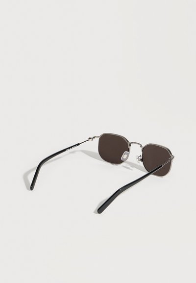 Κομψά γυαλιά ηλίου με μεταλλικό σκελετό, σκούρους ορθογώνιους φακούς και μαύρα κυρτά μπράτσα που ακουμπούν σε μια επίπεδη επιφάνεια.