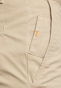 Beige cargobyxor med en sidoficka med söm och en liten orange etikett. Tyget är slätt med regular fit.