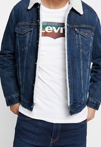 Man som bär en mörkblå jeansjacka med vitt fleecefoder över en vit T-shirt med flerfärgat randigt logo och mörkblå jeans.