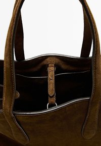 Grand sac fourre-tout en suède marron avec une texture douce, présentant une finition en cuir lisse et des poches intérieures. Comprend un clip en métal et des poignées robustes.