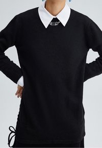 Maglione nero con maniche lunghe, polsini a costine e una camicia bianca col collarino sotto. Presenta un accento decorativo a catena sul colletto.