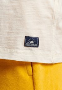 Jasno-kremowa tkanina koszulki z fakturowanym wykończeniem posiada granatową prostokątną metkę z grafiką wschodu słońca i napisem "SUPERDRY" na dolnym końcu.