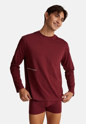 Junger Mann, der ein passendes bordeauxrotes Langarmshirt und Boxershorts trägt, lächelt und zieht den Saum des Shirts leicht nach vorne vor einem weißen Hintergrund.