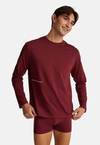 Bordová dlouhá rukávová košile s kulatým výstřihem a logem na textu, spárovaná s odpovídajícími bordovými těsnými šortkami. Materiál vypadá měkký a elastický.