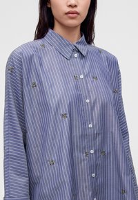 Femme portant une chemise bleue à boutons avec des rayures verticales blanches et de petits motifs de feuilles vertes brodées.
