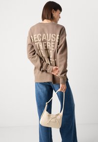 Brun sweatshirt med fet tekst på baksiden, kombinert med en lys beige skulderveske. Modellen har på seg blå vide jeans.