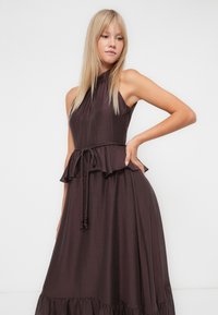 Dunkelbraunes ärmelloses Kleid mit hohem Ausschnitt, geraffter Taille und ausgestelltem Rock. Es hat eine Kordelzugdetails mit kleinen Verzierungen.