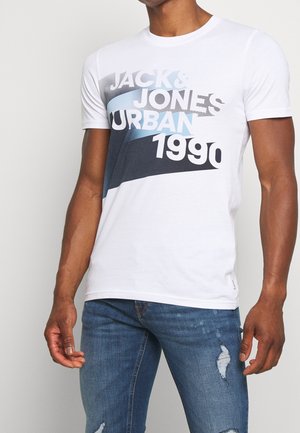 Homme portant un T-shirt blanc à manches courtes avec le texte "JACK & JONES URBAN 1990" et un jean bleu délavé, sur fond blanc.