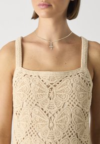 Beige heklete topp med sommerfugl-mønster og firkantet hals, med brede stropper. Sølvkjede med korsanheng.