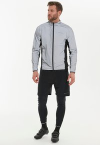 Lys grå vindjakke med sorte sidepaneler, lynlås lukning, båret over sorte shorts og leggings. Har refleksdetaljer og et sporty design.