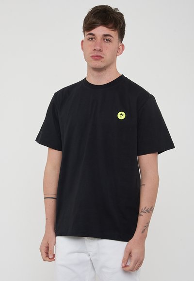 T-shirt en coton noir à manches courtes et col rond, avec un petit logo de smiley jaune sur la poitrine gauche.