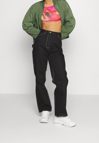 Grüne Bomberjacke, pinkes Cropped-Top und schwarze weite Hose mit weißem Nähen. Warum Sneaker runden das Outfit ab.