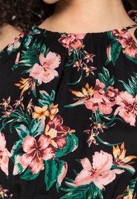 Nahaufnahme eines schwarzen Kleides mit einem tropischen Blumenmuster in Pink, Orange und Grün, das von einer Person mit lockigem Haar getragen wird.