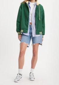 Chaqueta bomber verde con capucha amarilla, combinada con una camiseta a rayas azul y pantalones cortos de mezclilla desgastados. Zapatillas deportivas blancas completan el conjunto.