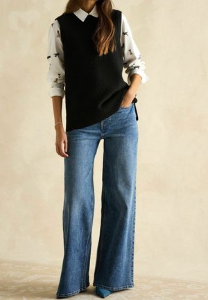 Femme portant un pull sans manches noir par-dessus une chemise blanche à motifs de chiens, assortie d'un jean bleu à jambes larges et de chaussures bleues pointues.