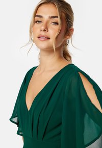 Zelené šifónové šaty s hlubokým véčkovým výstřihem, volnými rukávy a nařaseným pasem. Měkká textura a elegantní splývání jsou pozoruhodné vlastnosti.
