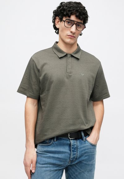 Polo vert olive avec un motif texturé, manches courtes et col. Associé à un jean bleu et une ceinture noire.