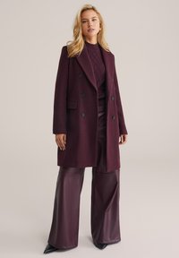 Burgundy, dupla gombos kabát texturált anyagból, nagy fekete gombokkal, magas derékú műbőr nadrággal és hegyes fekete sarkú cipővel párosítva.