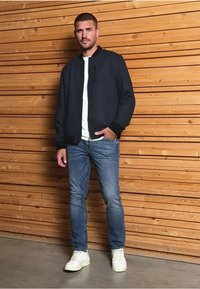 Navy bomber jacket over a white t-shirt, paired with blue jeans and white sneakers; standing against a wooden backdrop.