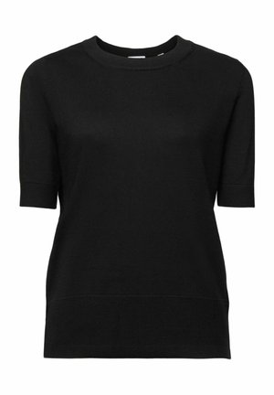 Kvinde iført sort t-shirt, sorte bukser med hvide geometriske mønstre og lys beige frakke mod en lyseblå baggrund.