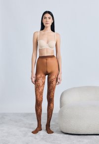 Καφέ αδιαφανές καλσόν με λουλουδένιο δαντελωτό μοτίβο, φορεμένο κάτω από ένα γυμνό σουτιέν, με ψηλή μέση και λεία υφή. Τοποθετημένο σε ανοιχτό φόντο.