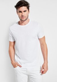 Valge puuvillane T-särk ümmarguse kaelusega ja lühikeste varrukatega, mille esiküljel on reljeefne "GUESS" logo. Mudel kannab heledat värvi pükse.