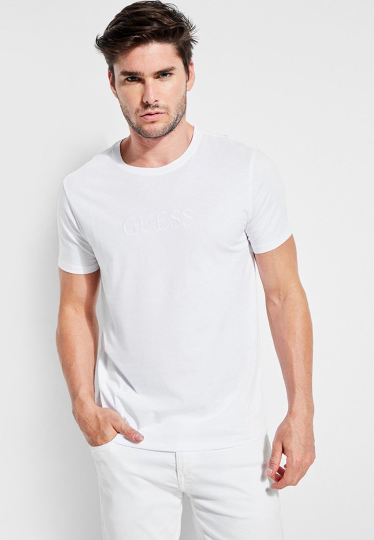 Valge puuvillane T-särk ümmarguse kaelusega ja lühikeste varrukatega, mille esiküljel on reljeefne "GUESS" logo. Mudel kannab heledat värvi pükse.