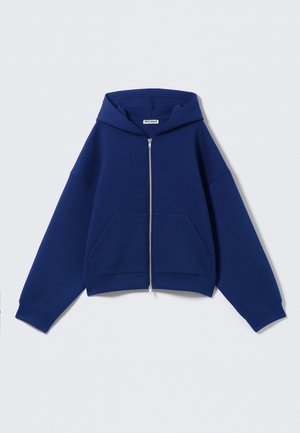Sweat à capuche bleu zippé avec poches avant, manches longues et une étiquette "Weekday" à l'intérieur du col.