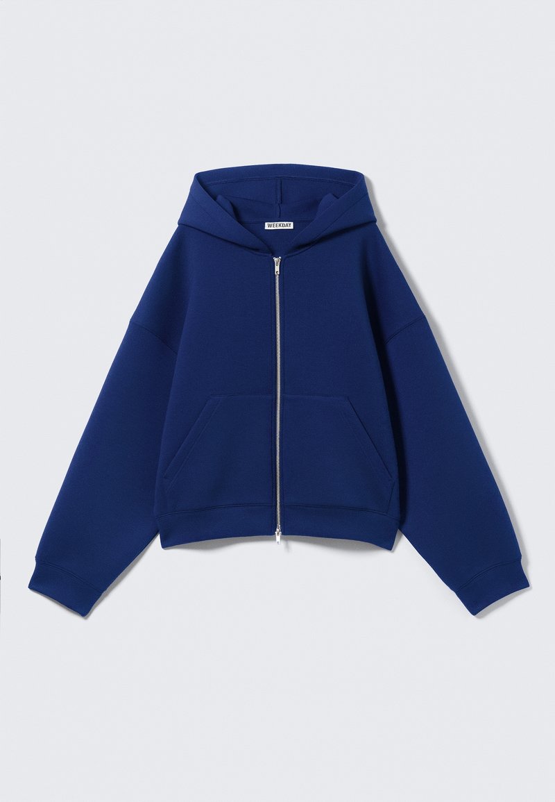 Sweat à capuche bleu zippé avec poches avant, manches longues et une étiquette "Weekday" à l'intérieur du col.