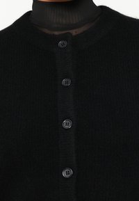 Black knitted cardigan featuring five black buttons, round neckline, and a soft textured fabric. Accent of a sheer black turtleneck underneath.