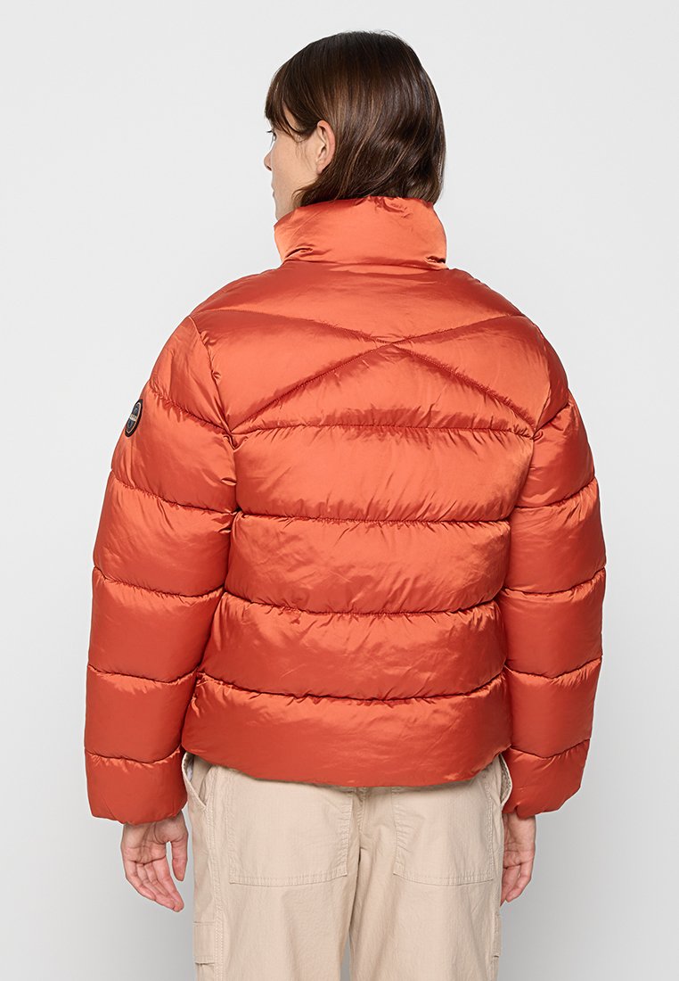 Person in orange Steppjacke mit hohem Kragen und beigen Hosen, von hinten vor einem schlichten Hintergrund betrachtet.