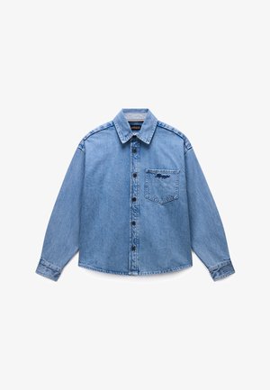 Camisa de manga larga de mezclilla azul claro, con botones, cuello y bolsillo frontal que presenta un pequeño detalle bordado oscuro.