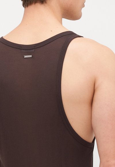 Débardeur côtelé marron foncé avec de larges emmanchures et un col cousu. Une petite étiquette en métal est visible à l'arrière.