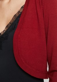 Close-up of a person wearing a black lace-trimmed top underneath a red knitted cardigan with rounded edges.