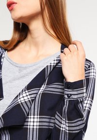 Rutigt blått och vitt skjorta med långa ärmar. Har en knäppt manschett och mjukt flanellmaterial. Bärs över en grå rundhalsad tröja.