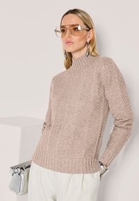 Frau trägt einen beigen Kabelstrick-Turtleneck-Pullover, übergroße, klare braune Sonnenbrille, silberne Ohrringe, ein Armband und hält eine kleine silberne Handtasche.