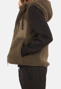 Chaleco de forro polar en verde oliva con capucha, que incluye un bolsillo en el pecho con cremallera y bolsillos laterales. Textura suave con mangas negras en contraste.