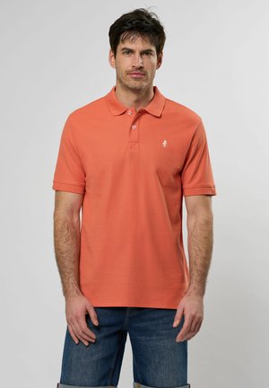 Chemise polo orange en tissu texturé, avec deux boutons au col et un petit logo blanc sur la poitrine gauche. Associée à un short en denim.