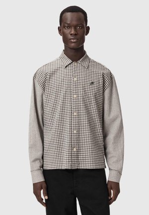 Mann trägt ein beige-braunes kariertes Hemd mit Knöpfen, gestreiften Ärmeln und schwarze Hose vor schlichtem grauem Hintergrund.