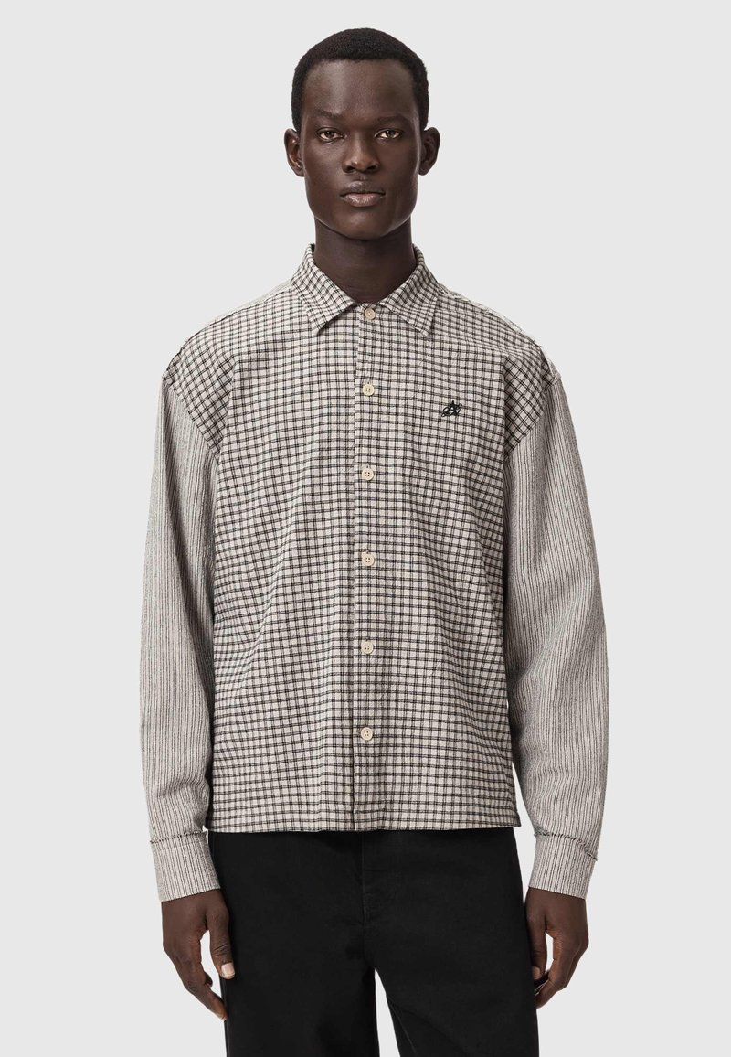 Homme portant une chemise à carreaux beige et marron à boutons avec des manches rayées et un pantalon noir, sur un fond gris uni.