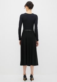 Un bodysuit negro de manga larga combinado con una falda midi plisada negra, que lleva un cinturón negro ajustado y zapatos de tacón negros. Textura de tela suave.