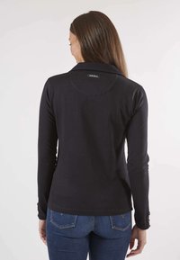 Chemise noire à manches longues avec col, présentant un empiècement rond et des détails de boutons sur les poignets. Le tissu semble lisse et ajusté.