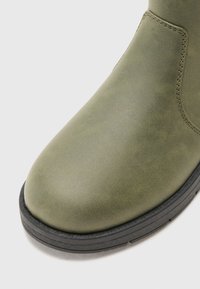 Bota de cuero verde oliva con punta redondeada, textura suave y suela de goma negra en contraste. Presenta un sutil detalle de costura.