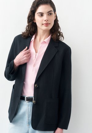 Młoda kobieta ubrana w czarną marynarkę na jasnoróżowej koszuli z kołnierzykiem i jasnoniebieskich dżinsach, stojąca na białym tle.