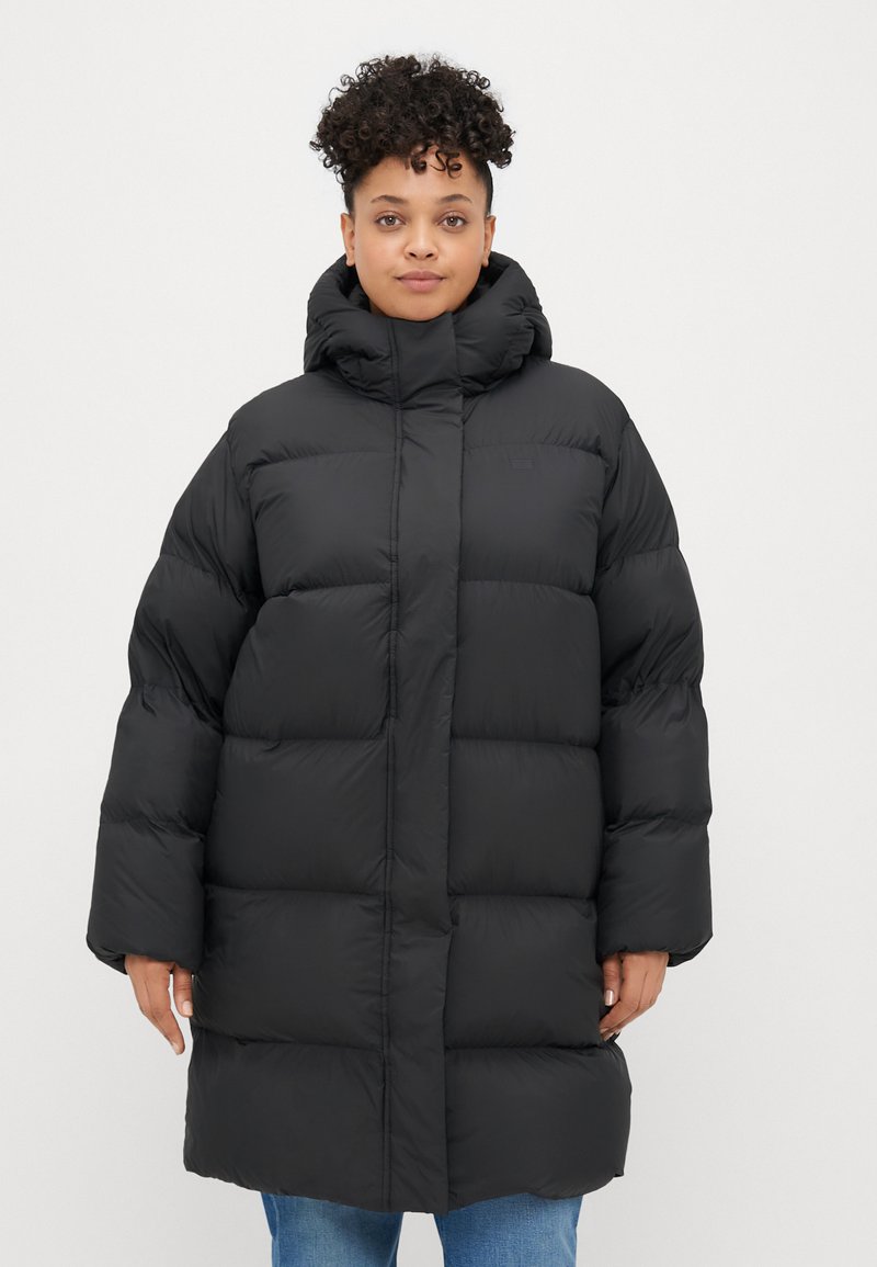 Black puffer coat with a high collar, featuring a boxy design, large quilted sections, and a front zipper. Textured, insulated fabric.