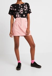 Kvinna i svart blommig kortärmad topp, rosa minikjol med stora fickor, rosa strumpor och svarta sneakers som står mot vit bakgrund.